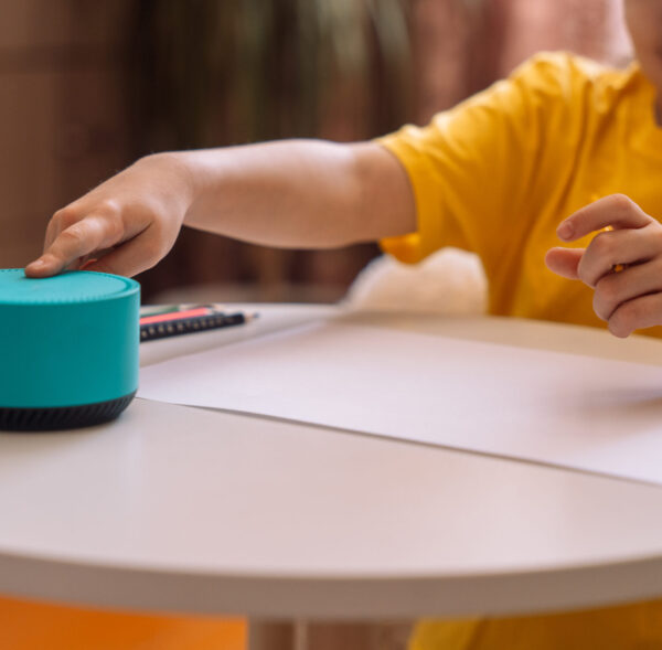 A child draws at the table and talks and presses on a blue smart speaker.